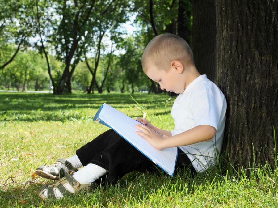 Dreng der sidder under et træ og læser i en bog